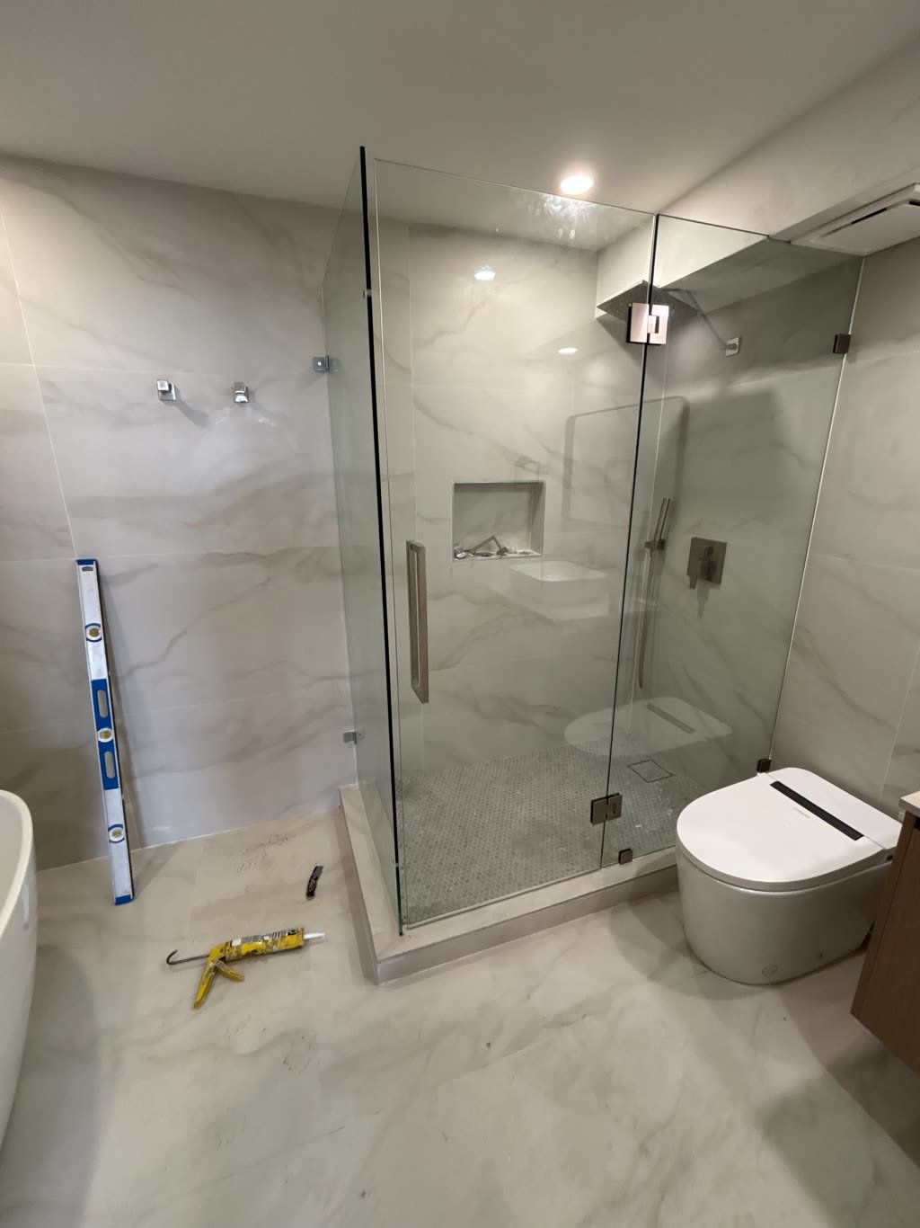 A bathroom with a corner shower enclosed in frameless glass, marble-look walls, a recessed shelf niche, a built-in corner bench and a mosaic tile floor.